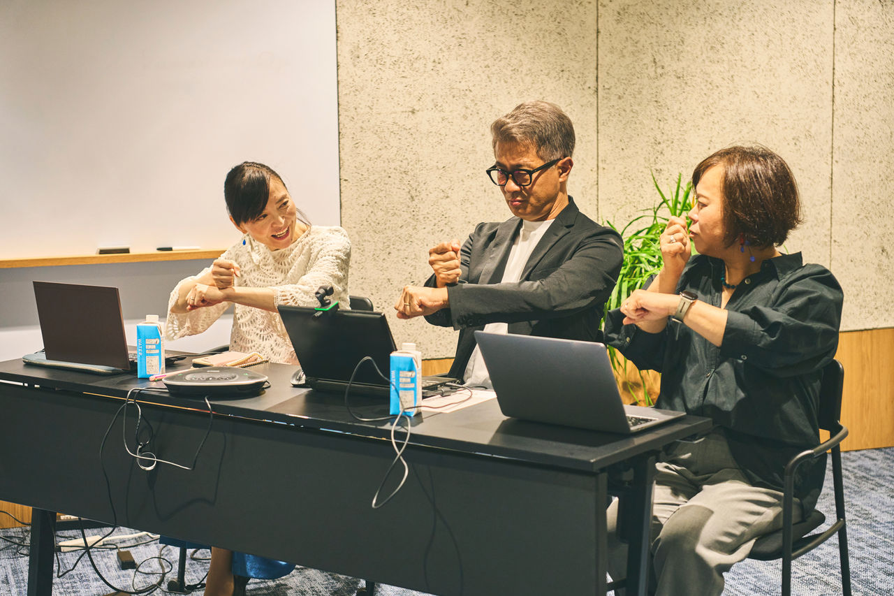 写真：笑顔で「お疲れさま」の手話を教える松森さんと、楽しそうに手話を教わる植木さんと千葉